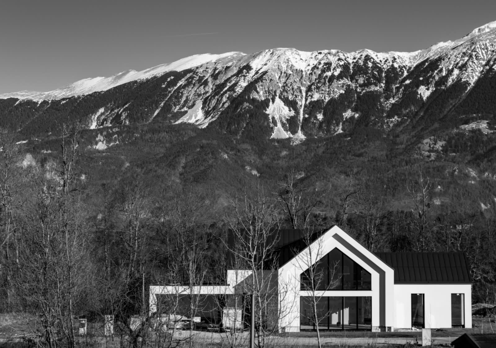 A modern gabled house at the foot of a snow-capped mountain range, in black and white.